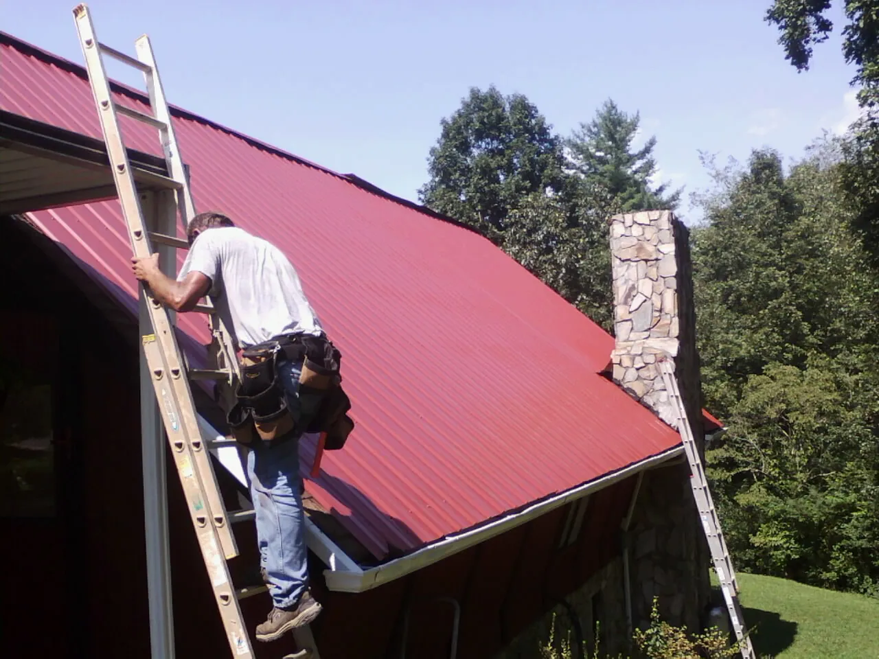 Roofer on a ladder beside a red metal roof and stone chimney — Soffit & Fascia Repair by Roofing Brothers in Woodsboro